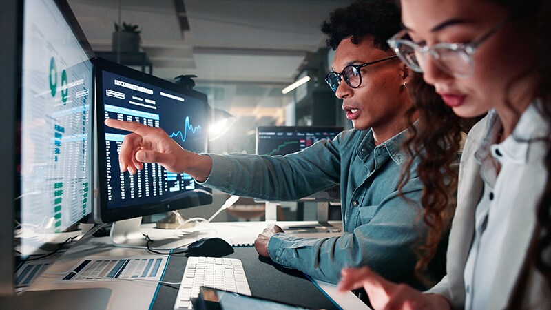 Two individuals in a modern office collaborate at computers displaying data. A young man points at his screen while a young woman focuses on a tablet.