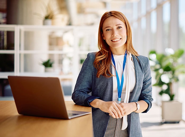 Professional woman in gray blazer standing in modern office with laptop, smiling at camera