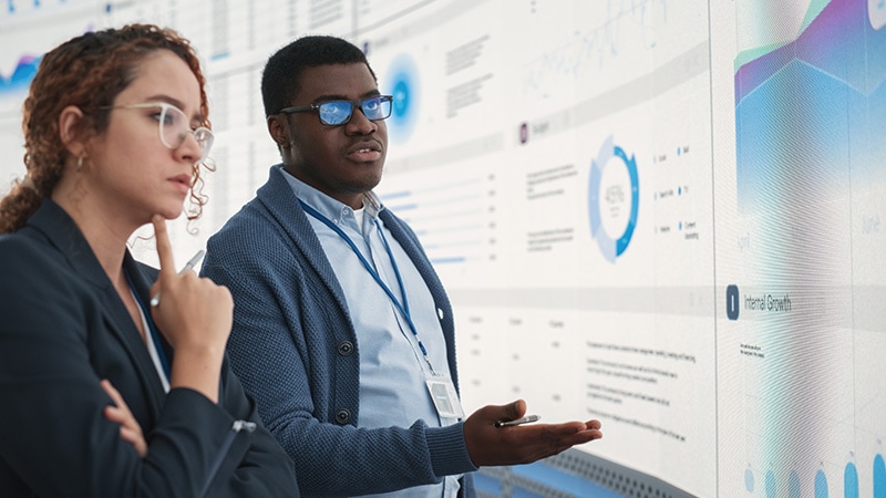 Two professionals discuss data in front of a large digital display filled with graphs and charts. One man gestures while the woman listens thoughtfully.