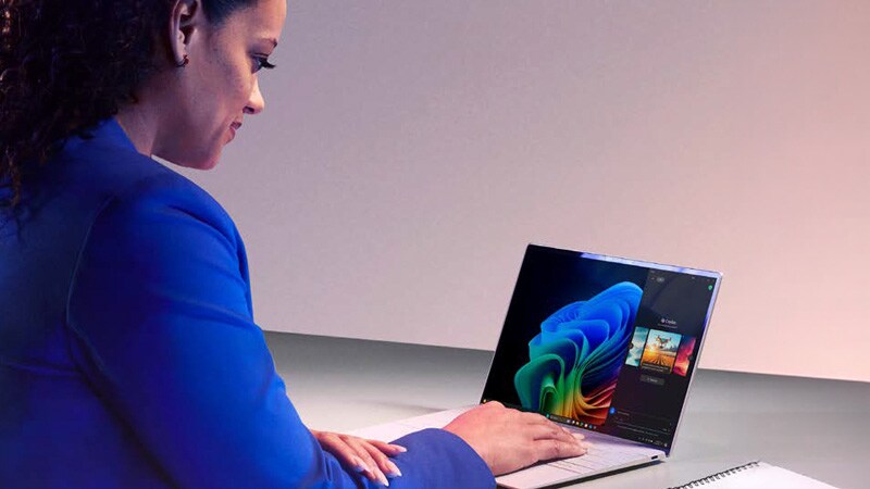 A woman in a blue blazer is sitting at a modern workstation, working on a colorful laptop.