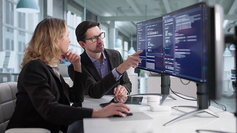 A man and a woman collaborate in a modern office, analyzing programming code displayed on computer screens.